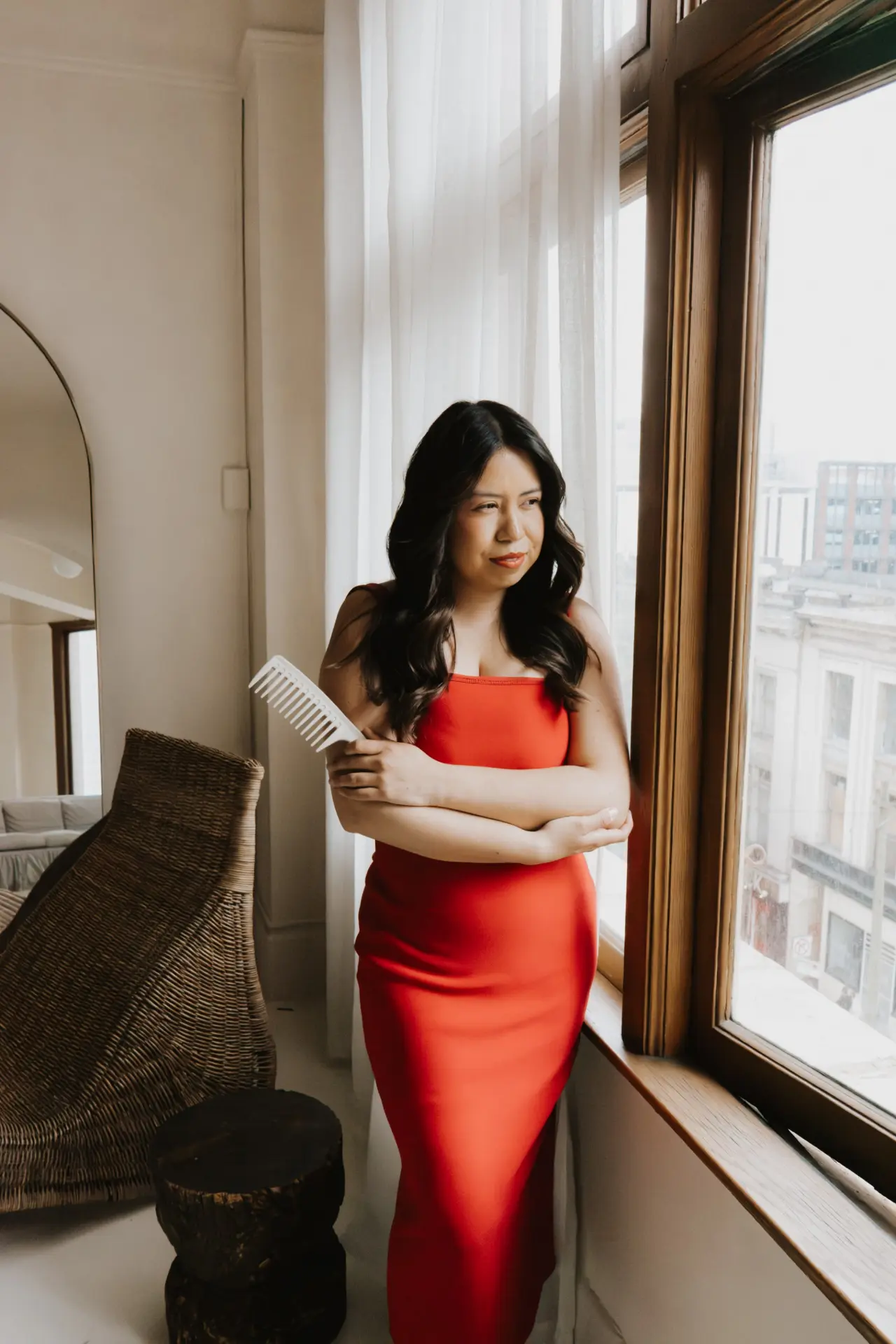 Rebeca in a red dress holding a wide-tooth comb beside a sunlit window